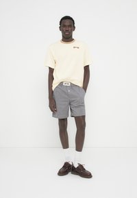 Άνδρας που φοράει ανοιχτό μπεζ κοντομάνικο πουκάμισο, καρό σορτς, λευκές κάλτσες και καφέ δερμάτινα παπούτσια, στέκεται μπροστά σε απλό λευκό φόντο.