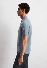 Jasnoniebieska koszulka polo z teksturowanej tkaniny, z krótkimi rękawami, z kołnierzykiem i zapięciem na dwa guziki, noszona z ciemnymi spodniami.