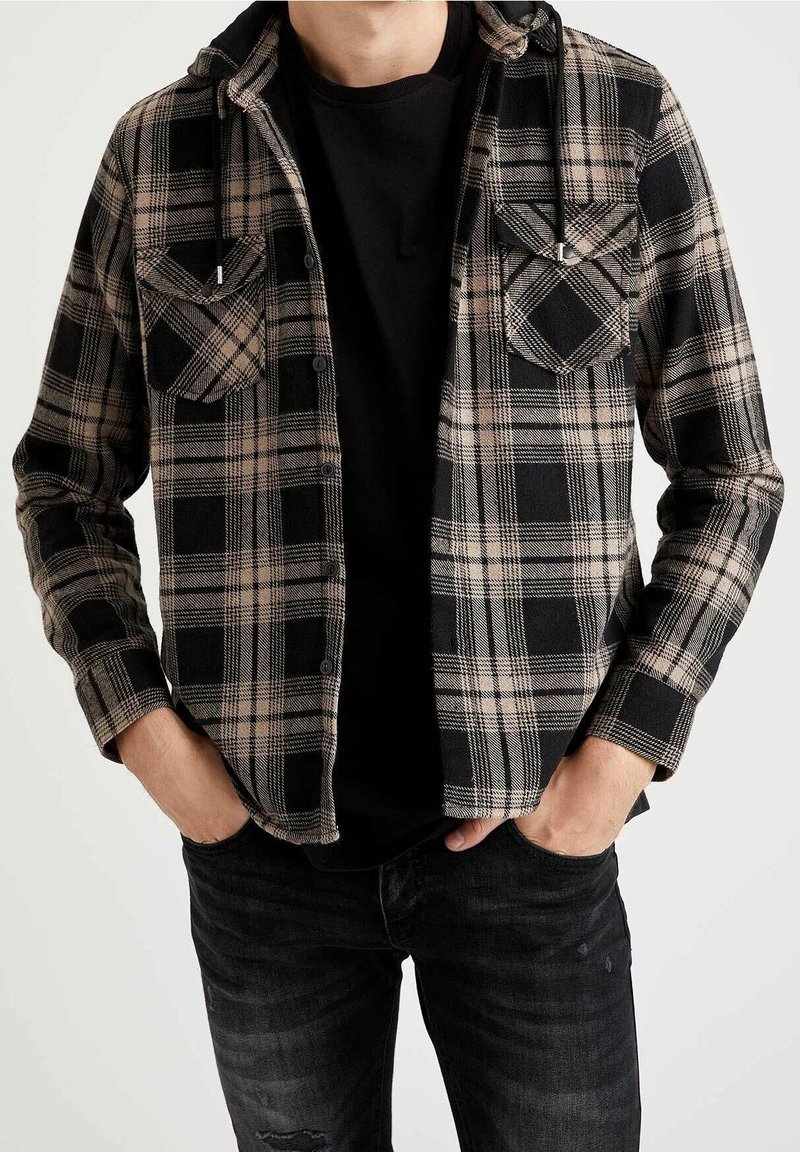 Homme portant une chemise à carreaux noire et beige boutonnée sur un T-shirt noir, les mains dans les poches d'un jean foncé sur fond blanc.