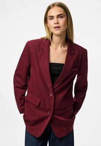 Vinstockfärgad blazer i slät tyg, med tvåknappsstängning, slagkrage och två framfickor, bärs över en svart topp.