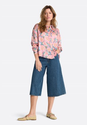 Frau steht und trägt ein langärmliges, rosa florales Hemd mit Knöpfen, breite blaue Jeans-Culottes und beige flache Schuhe vor einem weißen Hintergrund.