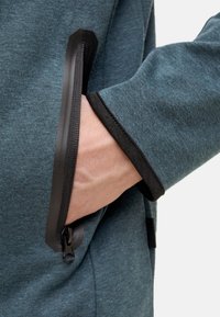 Teal kledingstuk met een ritszak, voorzien van zwarte rand en een zachte textuur. Hand geplaatst in de zak toont het ritsdetail en de nabijgelegen stof.