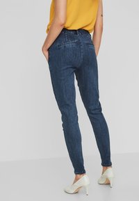 Jeans de mezclilla oscuros y ajustados con cintura alta, bolsillos traseros y un ligero estiramiento para mayor comodidad. Llevados con tacones blancos y una blusa amarillo mostaza.