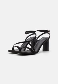 Sandales à talons en suede noir avec une conception à bout ouvert, présentant plusieurs fines lanières entrecroisées et un talon bloc. Bracelet de cheville ajustable inclus.