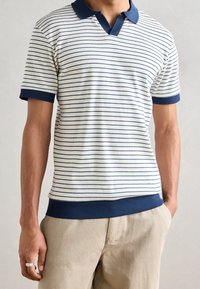 Bílo-námořnické pruhované polo tričko s límečkem, ripovanou texturou a námořnickými lemy na rukávech a spodním lemu. Nosené s béžovými kalhotami.