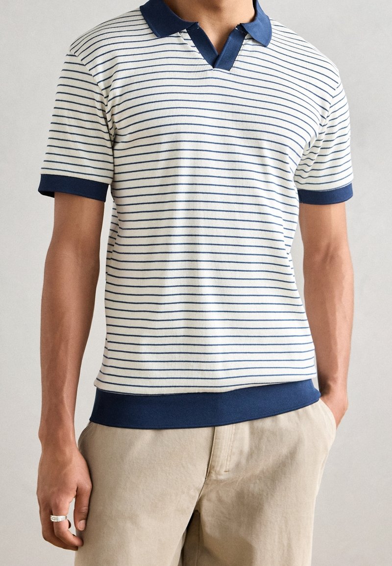 Bílo-námořnické pruhované polo tričko s límečkem, ripovanou texturou a námořnickými lemy na rukávech a spodním lemu. Nosené s béžovými kalhotami.
