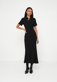 Femeie purtând o rochie midi neagră cu mâneci scurte, detaliu cu nod la gât, ciorapi negri și pantofi negri cu toc ascuțit, cu bretele și catarame.