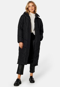 Sort lang pufferjakke med hætte, med quiltet mønster, lynlås og trykknaplukning, båret over en hvid sweater og sorte læderbukser.