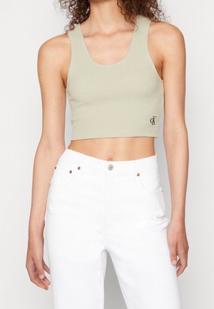 Kvinde iført lys grøn cropped tanktop med CK-logo og højtaljede hvide jeans, stående mod en ensfarvet baggrund.
