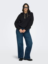 Nainen, jolla on musta huppari, jossa on kultainen sydän ja nyörit, tummansiniset leveälahkeiset farkut, mustat kengät ja mustat kissansilmälasit, seisoo käsivarret ristissä.