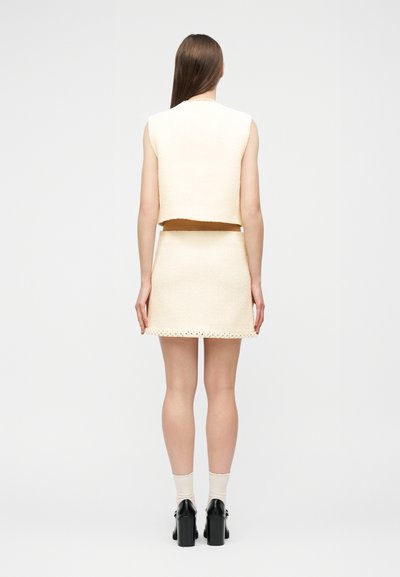 Γυναίκα με μακριά καστανά μαλλιά, φοράει αμάνικο κρεμ crop top και mini φούστα στον ίδιο χρωματισμό, λευκές κάλτσες και μαύρα παπούτσια με πλατφόρμα, όρθια και γυρισμένη με την πλάτη.