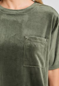 Primo piano di una camicia a maniche corte in tessuto verde vellutato con una tasca quadrata sul lato sinistro del petto.