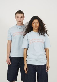 Light blue cotton t-shirts with short sleeves, featuring a curved pink "LEGACY STUDIOS" logo on the front. Worn with dark denim shorts.