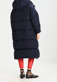 Námořnický modrý downový kabát s kapucí, se širokými vodorovnými pruhy, nošený přes červené legíny a černé boty s síťovanými punčochami.