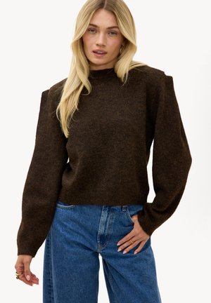 Femme blonde portant un pull marron foncé à manches longues avec des épaules rembourrées et un jean bleu taille haute, posant avec une main dans la poche.