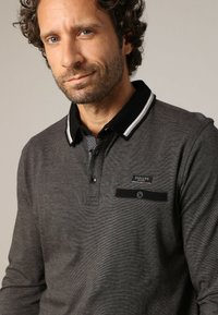 Donkergrijze long-sleeve polo met een geribbelde zwarte kraag. Heeft een knoopsluiting en een borstzak met een zwarte band en knoopdetail.