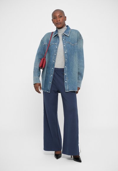 Staande persoon in een oversized denim jack, een grijze coltrui, marineblauwe wijde broek met zijknopen, zwarte hakken en een kleine rode schoudertas.