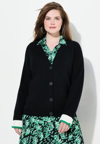 Cardigan noir à côtes avec cinq boutons et des poignets à volants contrastants blancs et verts, superposé sur une robe à motif floral vert.
