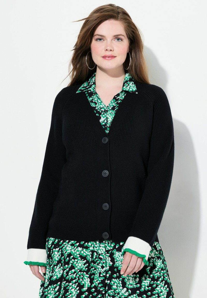 Cardigan noir à côtes avec cinq boutons et des poignets à volants contrastants blancs et verts, superposé sur une robe à motif floral vert.