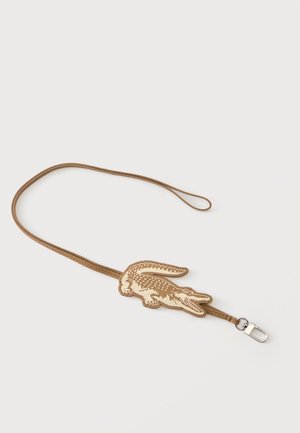 Lanière marron avec un patch en forme de crocodile beige et fauve ainsi qu'une attache métallique argentée sur fond blanc.