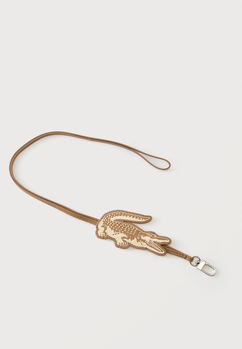 Lanière marron avec un patch en forme de crocodile beige et fauve ainsi qu'une attache métallique argentée sur fond blanc.
