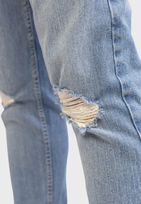 Svijetloplave traperice s istrošenim zakrpama na koljenima, s razderanim rubovima i izloženim bijelim nitima, koje imaju uski kroj.