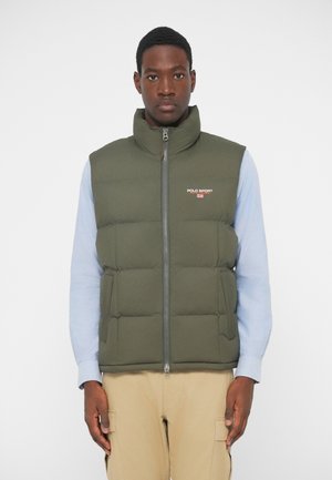 Zaļš pūšļu vests ar augstu apkakli, priekšējo rāvējslēdzēju un logo detaļu. Valkāts virs gaiši zilas garām piedurknēm krekla ar redzamām šuvēm.