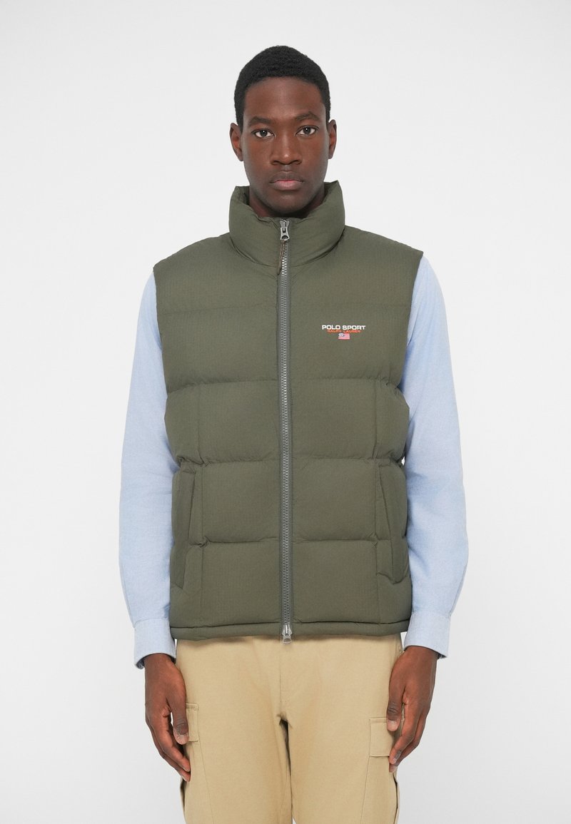 Zeleno pufferska brezrokavnica z visokim ovratnikom, sprednjim zadrgo in detajlom logotipa. Nosi se čez svetlo modro majico s dolgimi rokavi, ki ima vidne šive.