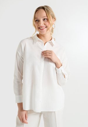 Chemise blanche à boutons en tissu léger, avec un col classique, des manches longues et une poche unique sur le côté gauche.