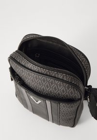 Sacoche grise compacte à bandoulière avec motif géométrique, poche zippée à l'avant et compartiment principal partiellement ouvert sur fond blanc.