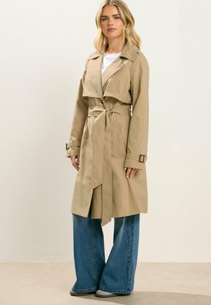 Lyshåret kvinde iført beige trenchcoat med bælte over hvid top med vide blå jeans og grå sko, stående mod en ensfarvet baggrund.