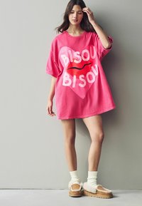T-shirt oversize rosa con il testo "BISOU" e un grafico a cuore rosso. La modella indossa calze di colore crema e scarpe beige con dettagli in pelliccia, in piedi contro un muro.
