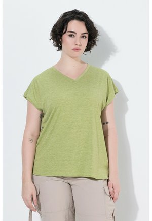 Kvinde med kort krøllet mørkt hår iført lysegrøn T-shirt med V-udskæring og beige cargobukser, stående op ad en hvid væg.