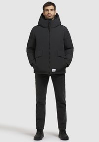 Sort polstret jakke med høj krave, frontlynlås og store sidelommer. Båret med mørke jeans og sorte støvler. Enkel design, ingen synlige mønstre.