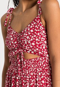 Röd blommig crop-top med justerbara knytband, utskärning och rynkad elastisk midja, prydd med vita blommor och gröna blad.