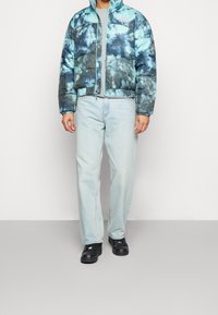 Hombre vistiendo una chaqueta de plumas de estampado tie-dye azul y negro, jeans de color azul claro, zapatos negros, de pie frente a un fondo blanco y gris liso.
