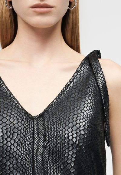 Kobieta ubrana w czarny, błyszczący bezrękawnik o fakturze skóry węża z cienkimi ramiączkami i srebrne kolczyki koła.