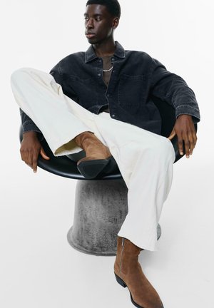 Camisetta di jeans nera con due tasche frontali, pantaloni bianchi a gamba larga e stivaletti alla caviglia in suede marrone, seduta su una sedia rotonda nera.
