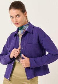 Paarse fleece jacket met een kraag, drukknoopjes en twee voorzakken. Zachte textuur, cropped ontwerp, gedragen over een bedrukt topje.