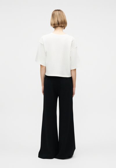 Άτομο με κοντά ξανθά μαλλιά που φοράει λευκή άνετη μπλούζα με κοντά μανίκια και μαύρο παντελόνι φαρδιάς γραμμής, στέκεται μπροστά σε απλό φωτεινό φόντο.