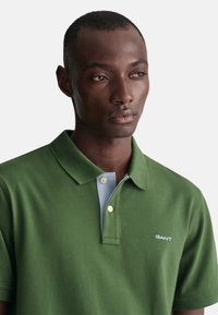 Grön polo t-shirt i bomull, med en ljusblå krage och tre knappar. T-shirten har en liten vit logotyp på bröstet.