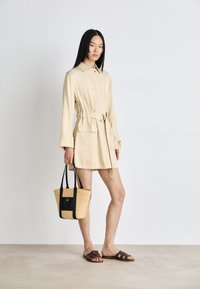 Béžové šaty s opaskom a dlhými rukávmi, so stredne veľkými vreckami, nosené s hnedými sklzmi. Drží prútenú tašku s čiernymi akcentmi. Hladká textura.