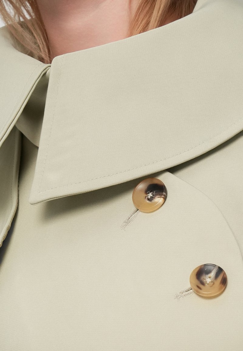 Détail d'un blazer vert clair avec un large col et deux grands boutons marron, présentant une finition en tissu texturé et des coutures soignées.