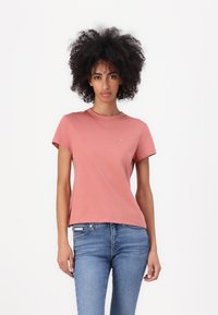 Trumpas rankovės, apvalaus iškirpimo t-shirt koralinės rožinės spalvos, pagamintas iš medvilnės, su mažu logotipu ant krūtinės ir laisvu kirpimu. Dėvimas su mėlynomis džinsomis.