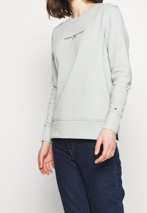 Sweatshirt gris clair avec des manches longues, des poignets en côtes et une taille en côtes. Présente un petit logo noir sur la poitrine. Assorti avec un jean bleu foncé.