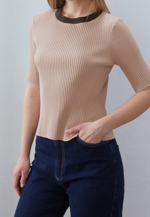 Femme portant un haut beige côtelé à manches courtes avec un col noir et un jean bleu foncé, main dans la poche du jean, sur fond gris uni.