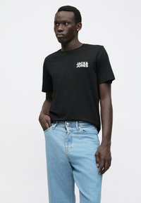 Vīrietis ģērbjas melnā "Jack & Jones" T-kreklā un gaiši zilos džinsos, stāvot ar vienu roku kabatā pret balto fona.