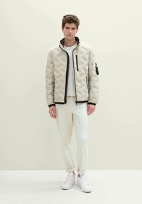 Helle beige Puffjacke mit einem gesteppten Muster, Reißverschluss vorne, seitlichen Taschen und schwarzen Akzenten. Getragen über einem grauen Pullover und weißen Hosen.