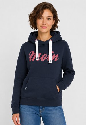 Jonge vrouw met kort krullend haar draagt een marineblauwe hoodie met roze "Moin" tekst en witte trekkoorden, gecombineerd met een blauwe spijkerbroek, staand tegen een witte achtergrond.