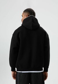 Zwarte oversized hoodie met capuchon en contrasterende stiksels langs de neklijn, gemaakt van zachte, gestructureerde stof. Licht losvallende pasvorm.